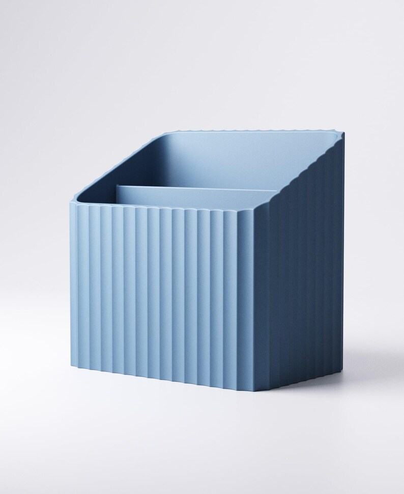 May include: A light blue desk organizer with a ribbed exterior design. The organizer has a slanted top and a divider inside, creating two compartments. The product is made of a solid material and is suitable for office or home use.