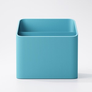 Puede incluir: Un organizador de escritorio azul turquesa con forma rectangular y textura acanalada vertical. El organizador tiene dos compartimentos internos y esquinas redondeadas. El fondo es blanco liso.