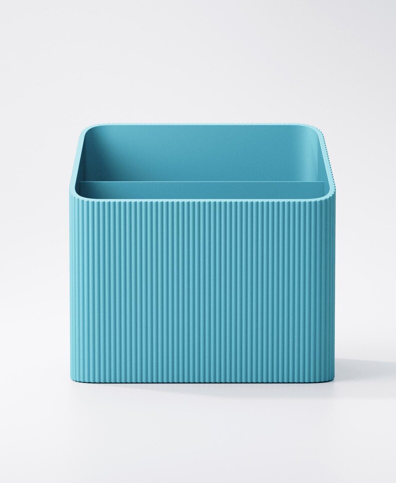 Puede incluir: Un organizador de escritorio azul turquesa con forma rectangular y textura acanalada vertical. El organizador tiene dos compartimentos internos y esquinas redondeadas. El fondo es blanco liso.