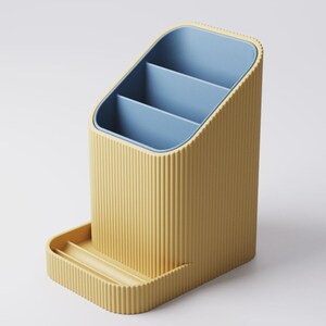 May include: A yellow desk organizer with a ribbed exterior and a blue interior. The organizer has three compartments and a small tray at the base. The design is modern and minimalist, suitable for office or home use.