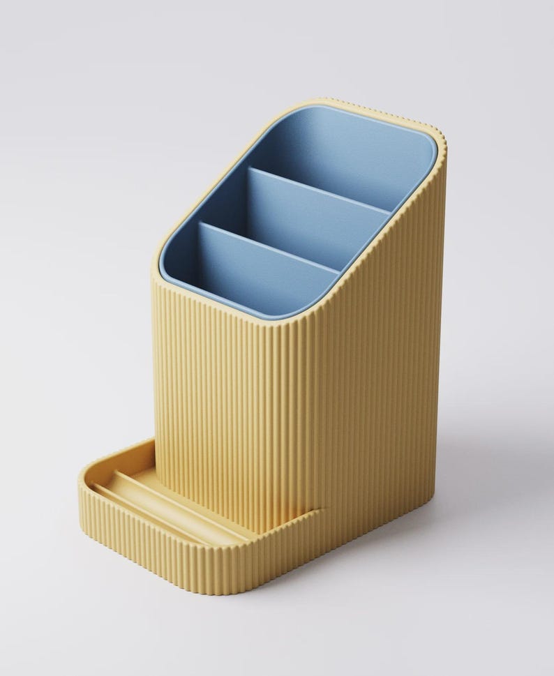 May include: A yellow desk organizer with a ribbed exterior and a blue interior. The organizer has three compartments and a small tray at the base. The design is modern and minimalist, suitable for office or home use.