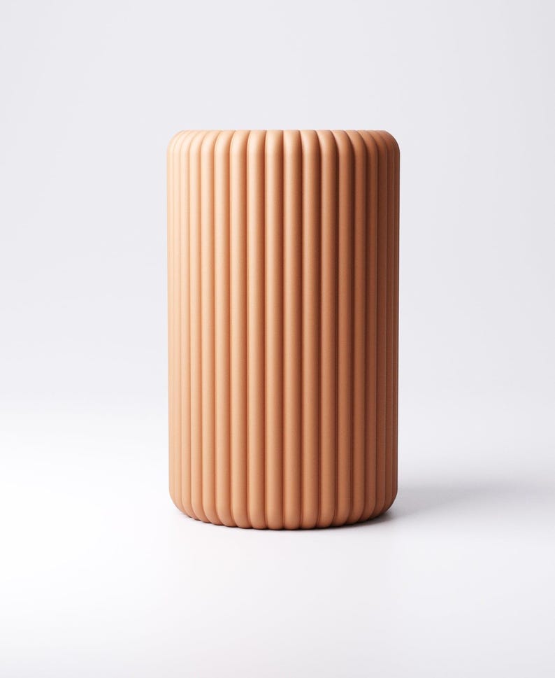 May include: A cylindrical, terracotta-colored vase with vertical, ridged detailing. The vase has a rounded top and base, and stands on a white background. The design is minimalist and modern.