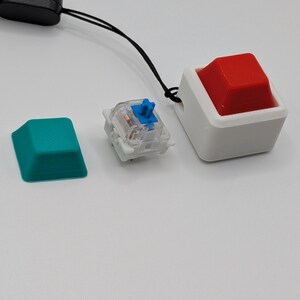 May include: An assortment of keyboard components, including a teal keycap, a clear mechanical switch with a blue stem, and a large red keycap in a white housing. A black lanyard is attached to the red keycap assembly.