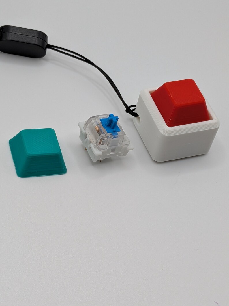 May include: An assortment of keyboard components, including a teal keycap, a clear mechanical switch with a blue stem, and a large red keycap in a white housing. A black lanyard is attached to the red keycap assembly.