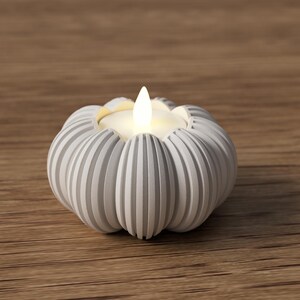 May include: A white, ribbed pumpkin-shaped candle holder with a lit, flickering candle. The candle holder is made of a light-colored material and sits on a wooden surface. The candle flame is a warm, inviting yellow.