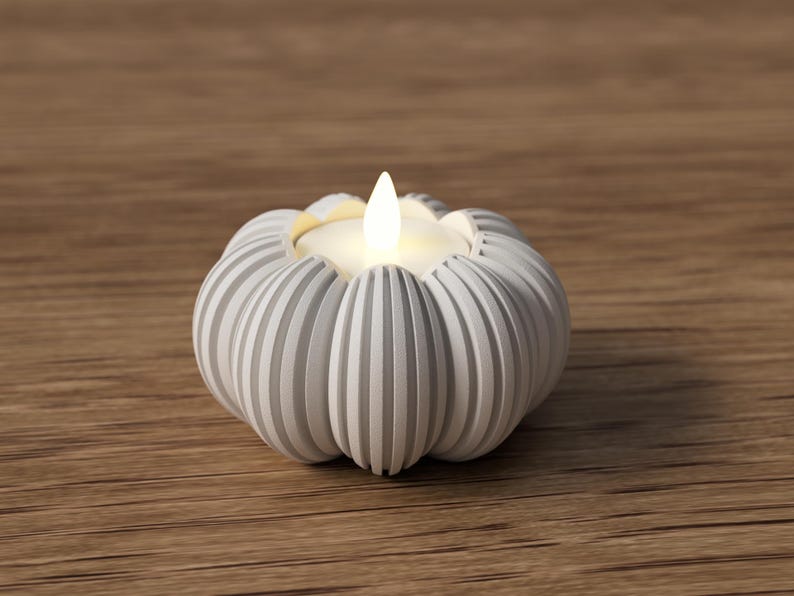 May include: A white, ribbed pumpkin-shaped candle holder with a lit, flickering candle. The candle holder is made of a light-colored material and sits on a wooden surface. The candle flame is a warm, inviting yellow.
