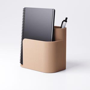 Pu&ograve; includere: Un organizer da scrivania beige con una texture a coste, contenente un taccuino nero a spirale e una penna argentata. L'organizer ha due scomparti, uno per il taccuino e uno per la penna.