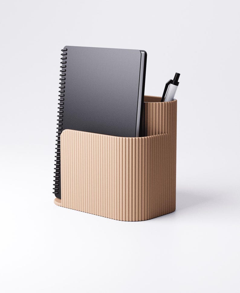 Pu&ograve; includere: Un organizer da scrivania beige con una texture a coste, contenente un taccuino nero a spirale e una penna argentata. L'organizer ha due scomparti, uno per il taccuino e uno per la penna.