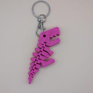 May include: A pink dinosaur skeleton keychain with a silver keyring and clasp. The dinosaur is made of connected pieces, with a detailed skull and spine. The keychain is a fun accessory.