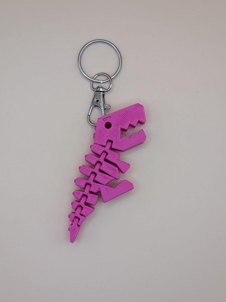 May include: A pink dinosaur skeleton keychain with a silver keyring and clasp. The dinosaur is made of connected pieces, with a detailed skull and spine. The keychain is a fun accessory.