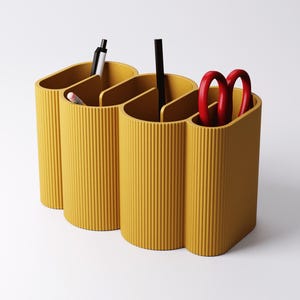 May include: Mustard yellow desk organizer with three connected compartments. The organizer has a ribbed texture and holds a pen, pencil, black pen, and red scissors. The design is modern and minimalist, suitable for office or home use.