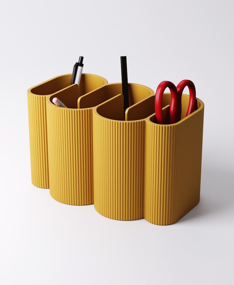 May include: Mustard yellow desk organizer with three connected compartments. The organizer has a ribbed texture and holds a pen, pencil, black pen, and red scissors. The design is modern and minimalist, suitable for office or home use.