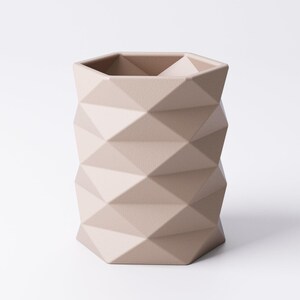 May include: A beige geometric vase with a faceted design. The vase has a hexagonal top and a textured surface. The neutral color and modern design make it a versatile decorative piece.