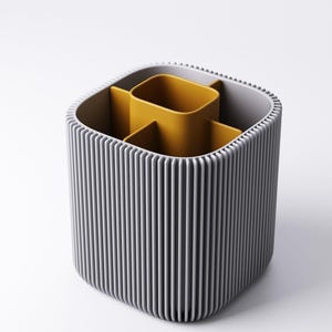 May include: A grey and yellow desk tidy with a ribbed exterior. The organiser has a square shape with rounded corners and contains several compartments. The interior compartments are yellow, and the exterior is grey.