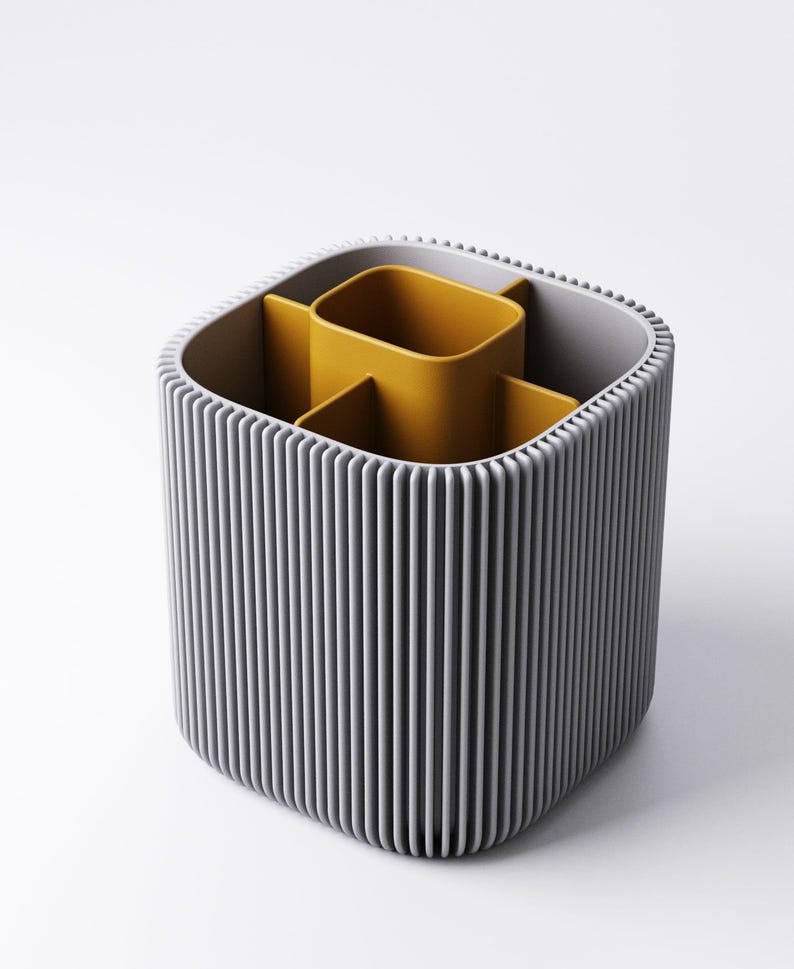 May include: A grey and yellow desk tidy with a ribbed exterior. The organiser has a square shape with rounded corners and contains several compartments. The interior compartments are yellow, and the exterior is grey.
