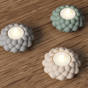 May include: Three decorative candle holders with lit candles. The holders are in shades of gray, green, and off-white, each with a textured, bubble-like design. The candles emit a warm glow, set against a wooden background.