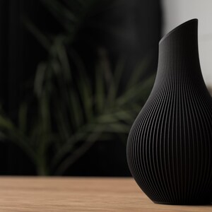 May include: A black vase with a unique, ribbed design. The vase has a curved, asymmetrical top and a rounded body. It is sitting on a wooden surface, with a blurred background of greenery.