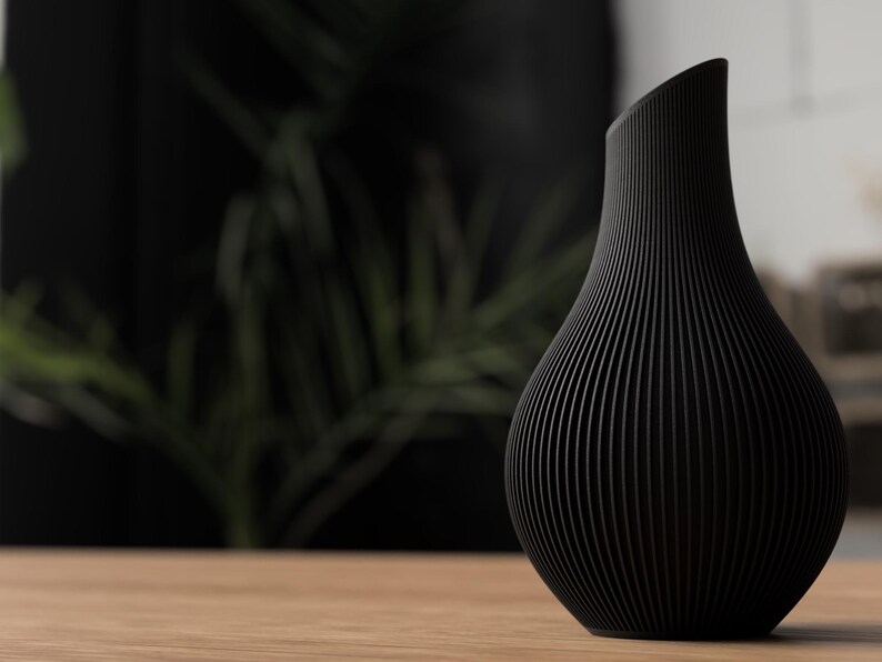May include: A black vase with a unique, ribbed design. The vase has a curved, asymmetrical top and a rounded body. It is sitting on a wooden surface, with a blurred background of greenery.