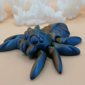 Op de afbeelding: Een gedetailleerd, iriserend blauw en bronzen spinnenbeeldje met acht poten en een getextureerd lichaam. De spin is het middelpunt, tegen een zachte, neutrale achtergrond met witte, kristallijne formaties.
