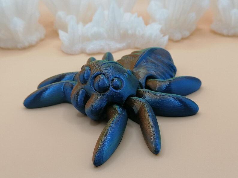 Op de afbeelding: Een gedetailleerd, iriserend blauw en bronzen spinnenbeeldje met acht poten en een getextureerd lichaam. De spin is het middelpunt, tegen een zachte, neutrale achtergrond met witte, kristallijne formaties.