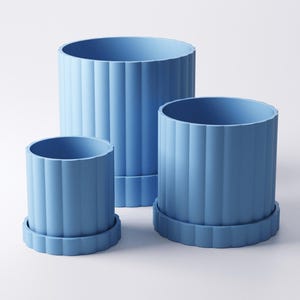 May include: Three light blue cylindrical planters of varying sizes. Each planter has vertical ridged detailing and sits on a matching round base. The planters are empty and ready for plants. The background is plain white.