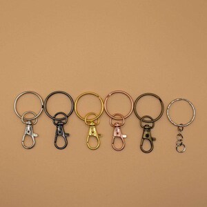 May include: A collection of keychains in various metallic finishes, including silver, black, gold, and rose gold. Each keychain features a round ring and a lobster clasp for easy attachment. The keychains are arranged in a row against a neutral background.
