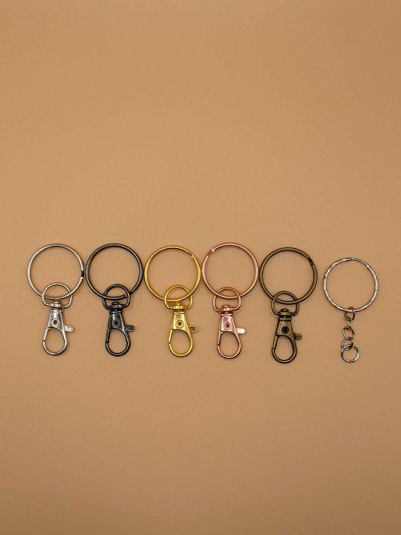May include: A collection of keychains in various metallic finishes, including silver, black, gold, and rose gold. Each keychain features a round ring and a lobster clasp for easy attachment. The keychains are arranged in a row against a neutral background.