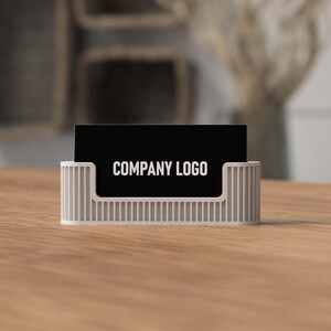 May include: A white business card holder with vertical lines holds a black business card. The card displays the text "COMPANY LOGO" in white. The holder sits on a wooden surface.