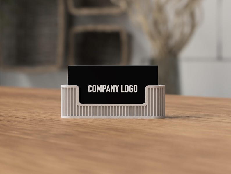 May include: A white business card holder with vertical lines holds a black business card. The card displays the text "COMPANY LOGO" in white. The holder sits on a wooden surface.