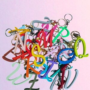 May include: A collection of colorful letter keychains in various fonts and hues, including red, yellow, green, and blue. Each letter is attached to a metal keyring. The keychains are scattered against a soft pink background.