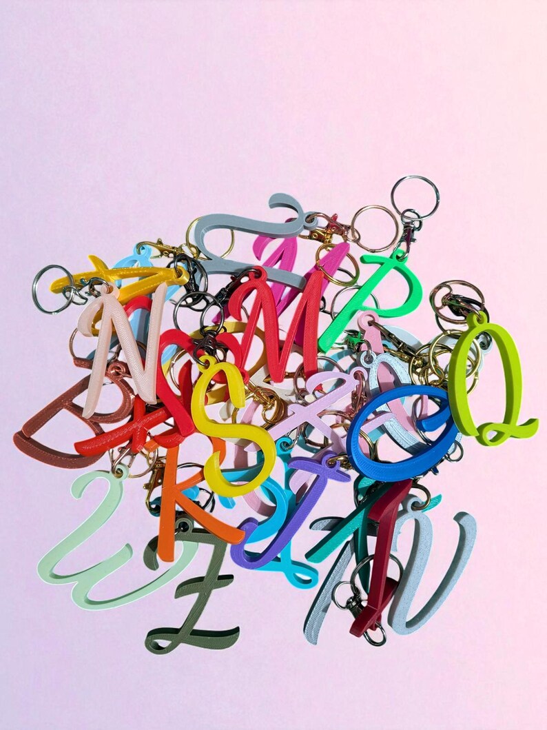 May include: A collection of colorful letter keychains in various fonts and hues, including red, yellow, green, and blue. Each letter is attached to a metal keyring. The keychains are scattered against a soft pink background.