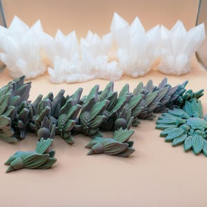 May include: A collection of 3D-printed fantasy sculptures. The pieces include white crystal formations and a segmented, green and purple dragon-like creature. A teal floral design is also present, all on a neutral background.