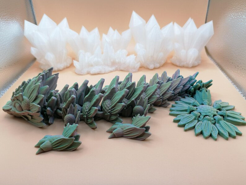 May include: A collection of 3D-printed fantasy sculptures. The pieces include white crystal formations and a segmented, green and purple dragon-like creature. A teal floral design is also present, all on a neutral background.