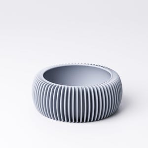 May include: A light gray, round bowl with vertical ribbed detailing on the exterior. The bowl has a smooth interior and a wide opening. The design is modern and minimalist, suitable for various uses.