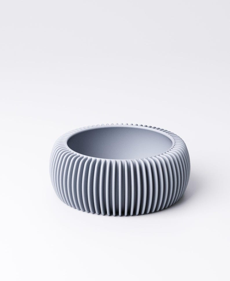 May include: A light gray, round bowl with vertical ribbed detailing on the exterior. The bowl has a smooth interior and a wide opening. The design is modern and minimalist, suitable for various uses.