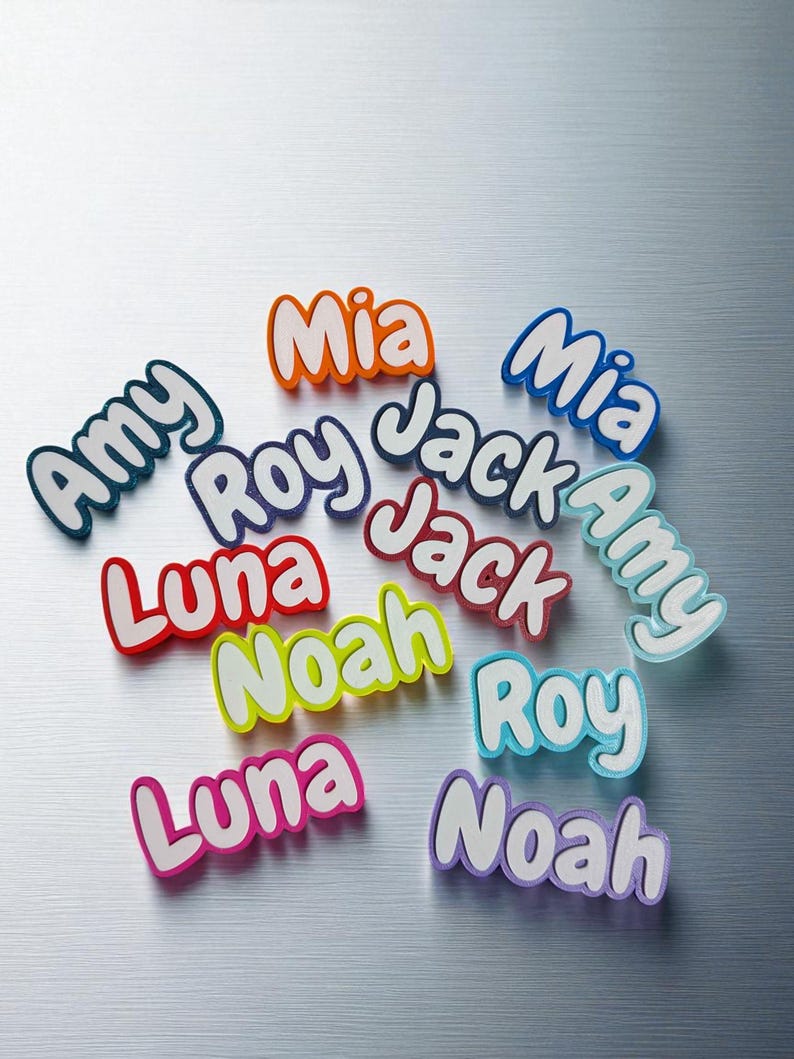 May include: A collection of colorful name cutouts, including "Amy," "Mia," "Jack," "Roy," "Luna," and "Noah." Each name is in a different color, with a white outline, and set against a metallic silver background.