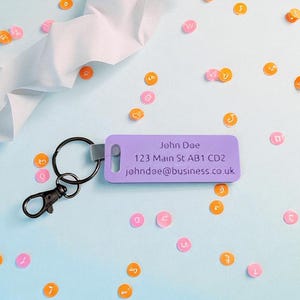 May include: A purple luggage tag with a black keyring and clasp. The tag has the text "John Doe", address, and email. Scattered around are orange and pink circular confetti with letters and numbers on a light blue background.