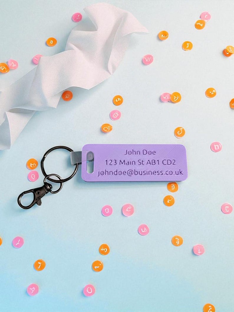 May include: A purple luggage tag with a black keyring and clasp. The tag has the text "John Doe", address, and email. Scattered around are orange and pink circular confetti with letters and numbers on a light blue background.