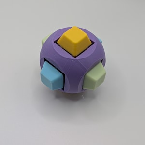 May include: A multi-colored puzzle cube with a spherical shape. The cube is primarily purple, with yellow, light blue, and green square protrusions. The puzzle is designed to be manipulated and solved.