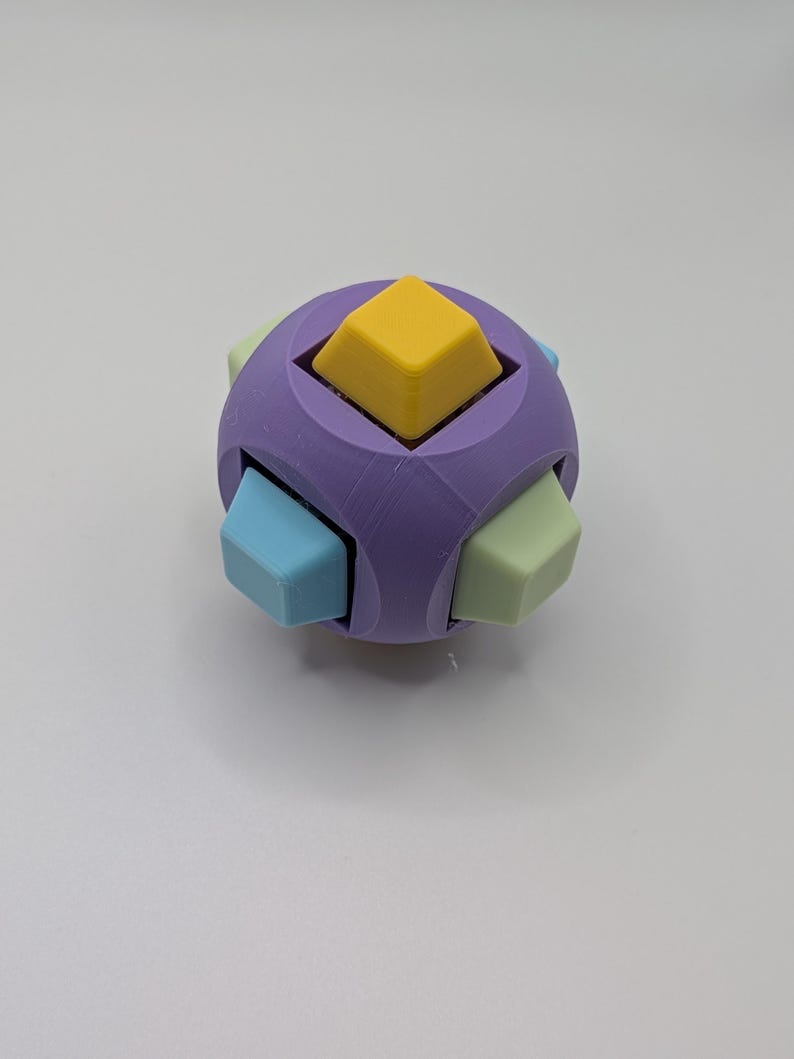 May include: A multi-colored puzzle cube with a spherical shape. The cube is primarily purple, with yellow, light blue, and green square protrusions. The puzzle is designed to be manipulated and solved.