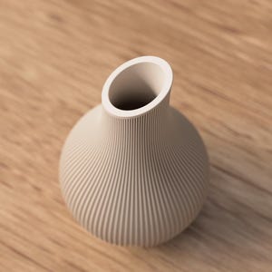 May include: A beige ceramic vase with a round body and a slanted opening. The vase has vertical, ridged detailing and sits on a wooden surface. The vase is a decorative home decor item.