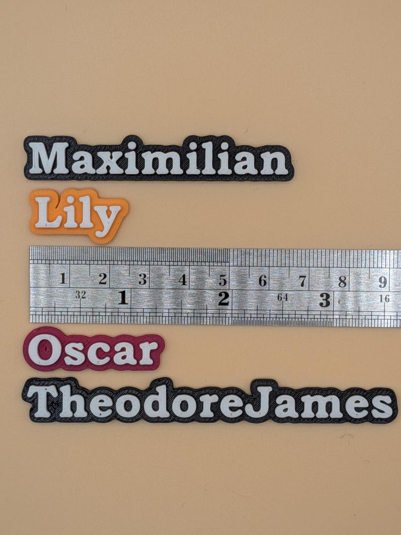 Może przedstawiać: Metalowa linijka z podziałką w centymetrach i kilka etykiet z imionami. Etykiety zawierają imiona "Maximilian", "Lily", "Oscar" i "TheodoreJames". Imiona są w białej czcionce z czarną obw&oacute;dką. "Lily" jest pomarańczowa.
