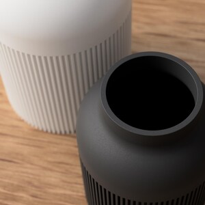 May include: Two modern vases, one white and one dark gray, sit on a wooden surface. The white vase is tall and cylindrical, with vertical ribbed detailing on the lower half. The dark gray vase is shorter and rounder, also with ribbed detailing.