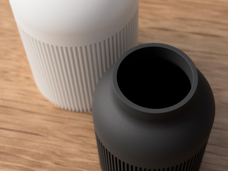 May include: Two modern vases, one white and one dark gray, sit on a wooden surface. The white vase is tall and cylindrical, with vertical ribbed detailing on the lower half. The dark gray vase is shorter and rounder, also with ribbed detailing.