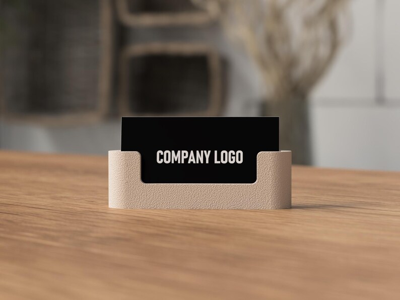May include: A business card holder with a black card displaying "COMPANY LOGO" in white text. The holder is a light beige color and sits on a wooden surface. The background is blurred, suggesting an office or workspace setting.