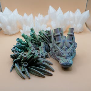 May include: A 3D-printed dragon sculpture with a teal and purple color scheme, featuring intricate details. The dragon is accompanied by several white, crystal-like structures in the background. The objects are displayed on a light tan surface.