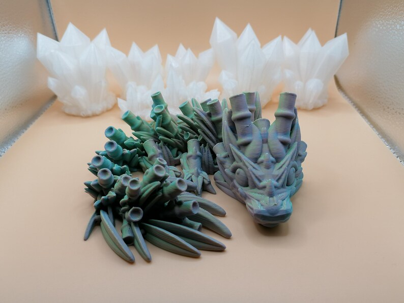 May include: A 3D-printed dragon sculpture with a teal and purple color scheme, featuring intricate details. The dragon is accompanied by several white, crystal-like structures in the background. The objects are displayed on a light tan surface.