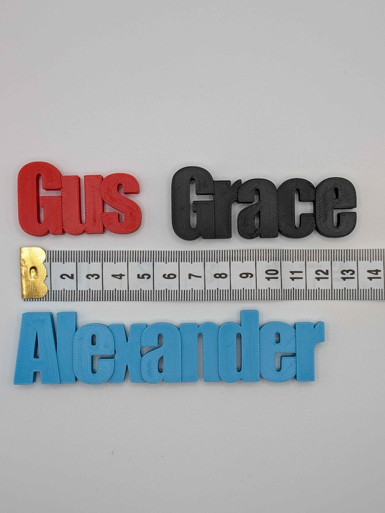 May include: Three 3D-printed names: "Gus" in red, "Grace" in black, and "Alexander" in blue. A ruler is placed next to the names for scale. The names are in a sans-serif font and appear to be made of plastic.