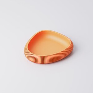 Puede incluir: Un cuenco naranja de forma triangular con un exterior acanalado. El cuenco tiene un interior liso y un dise&ntilde;o texturizado con l&iacute;neas verticales alrededor del borde. El cuenco est&aacute; hecho de un material s&oacute;lido y se encuentra sobre una superficie blanca.