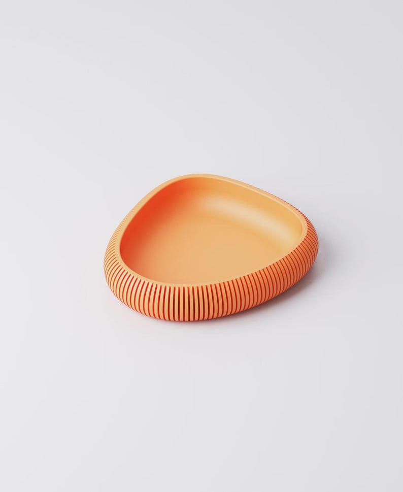 Puede incluir: Un cuenco naranja de forma triangular con un exterior acanalado. El cuenco tiene un interior liso y un dise&ntilde;o texturizado con l&iacute;neas verticales alrededor del borde. El cuenco est&aacute; hecho de un material s&oacute;lido y se encuentra sobre una superficie blanca.
